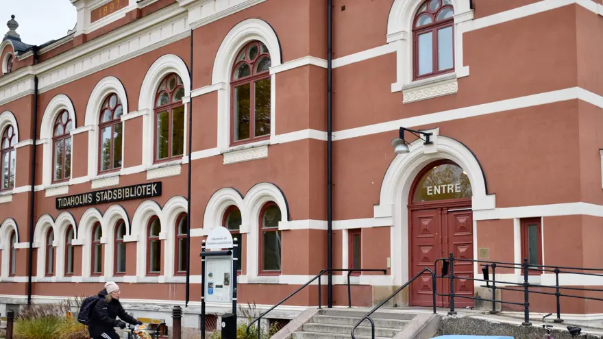 Tidaholms bibliotek, en röd byggnad med vita fönster som är formade som bågar längst upp. En kvinna cyklar förbi, på huset står skylten TIDAHOLMS BIBLIOTEK.