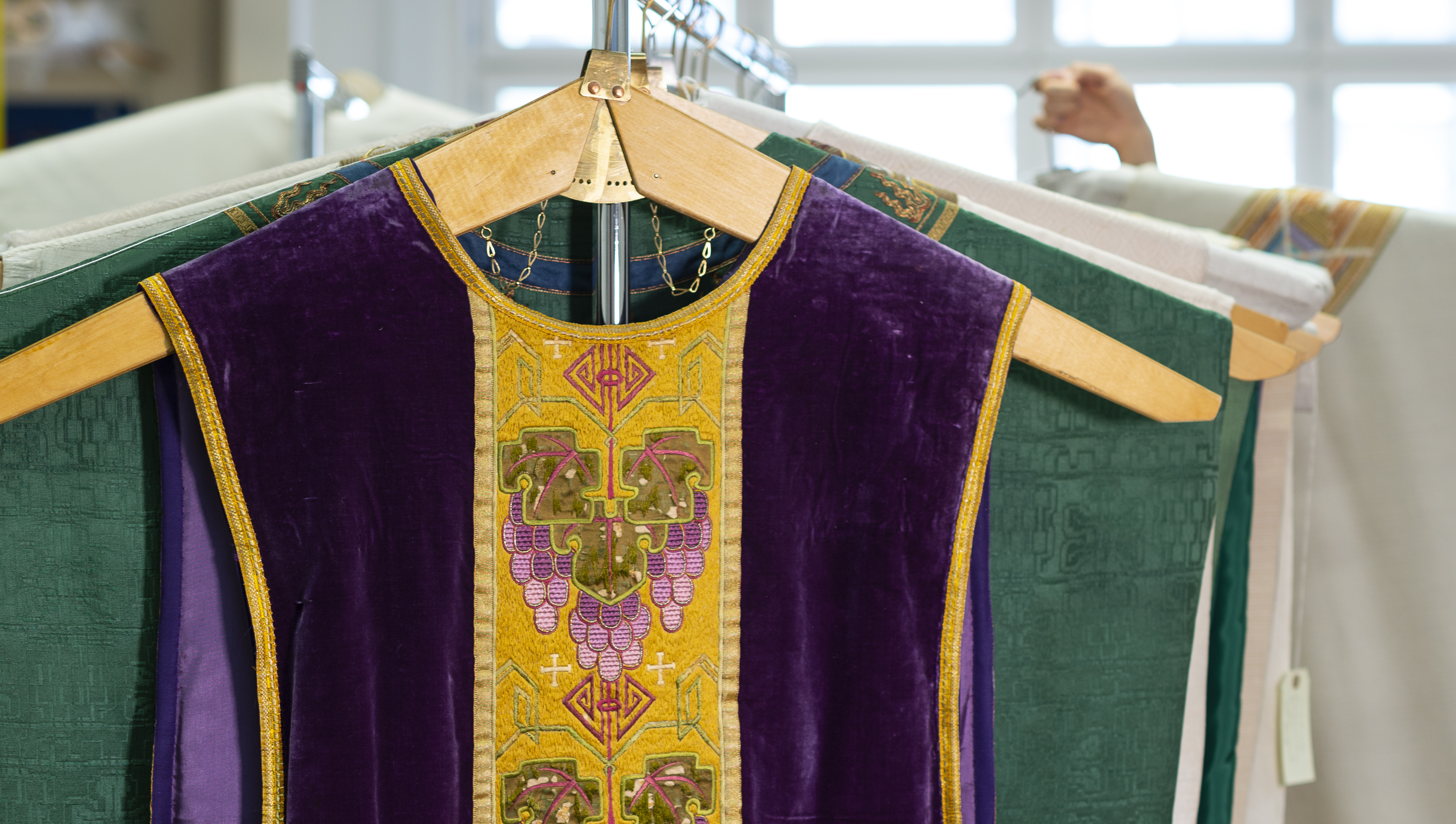 På en ställning hänger rader med mässhakar, längst fram en violett. I bakgrunden skymtar stora, ljusa fönster.