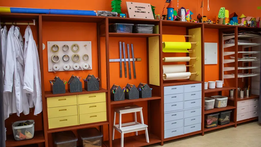 Ett hyllsystem fullt med pysselmaterial - pennor, linjaler, papper, tejp, plastlådor med tyger med mera. Till höger hänger vita labbrockar på en stång, teckningar ligger på tork på hyllor och längst upp står olika konstverk i papper. En vit träpall står längst ner. Väggen är målad orange och lådor är målade i ljust gult och blått.