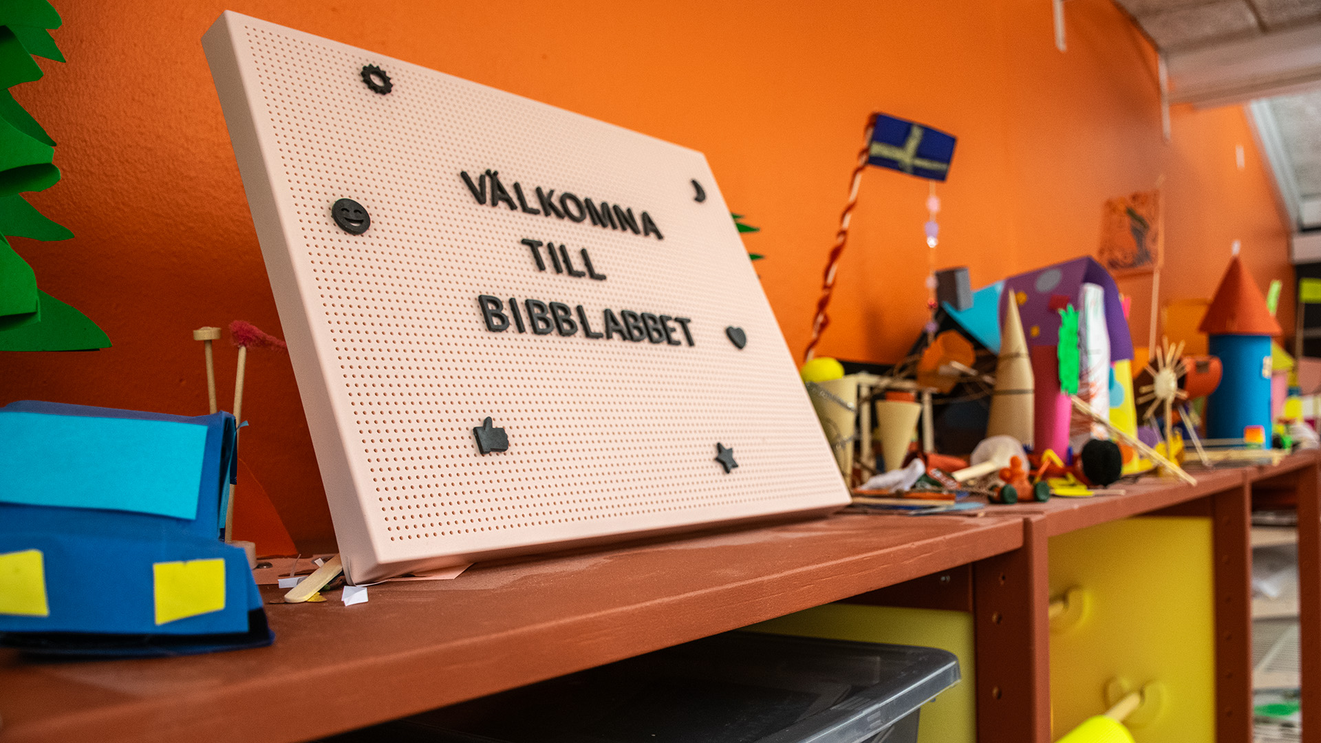 En skylt står längst upp på en hylla. På den står Välkomna till Bibblabbet med stora bokstäver. På den bruna hyllan finns olika skapelser i papper. 