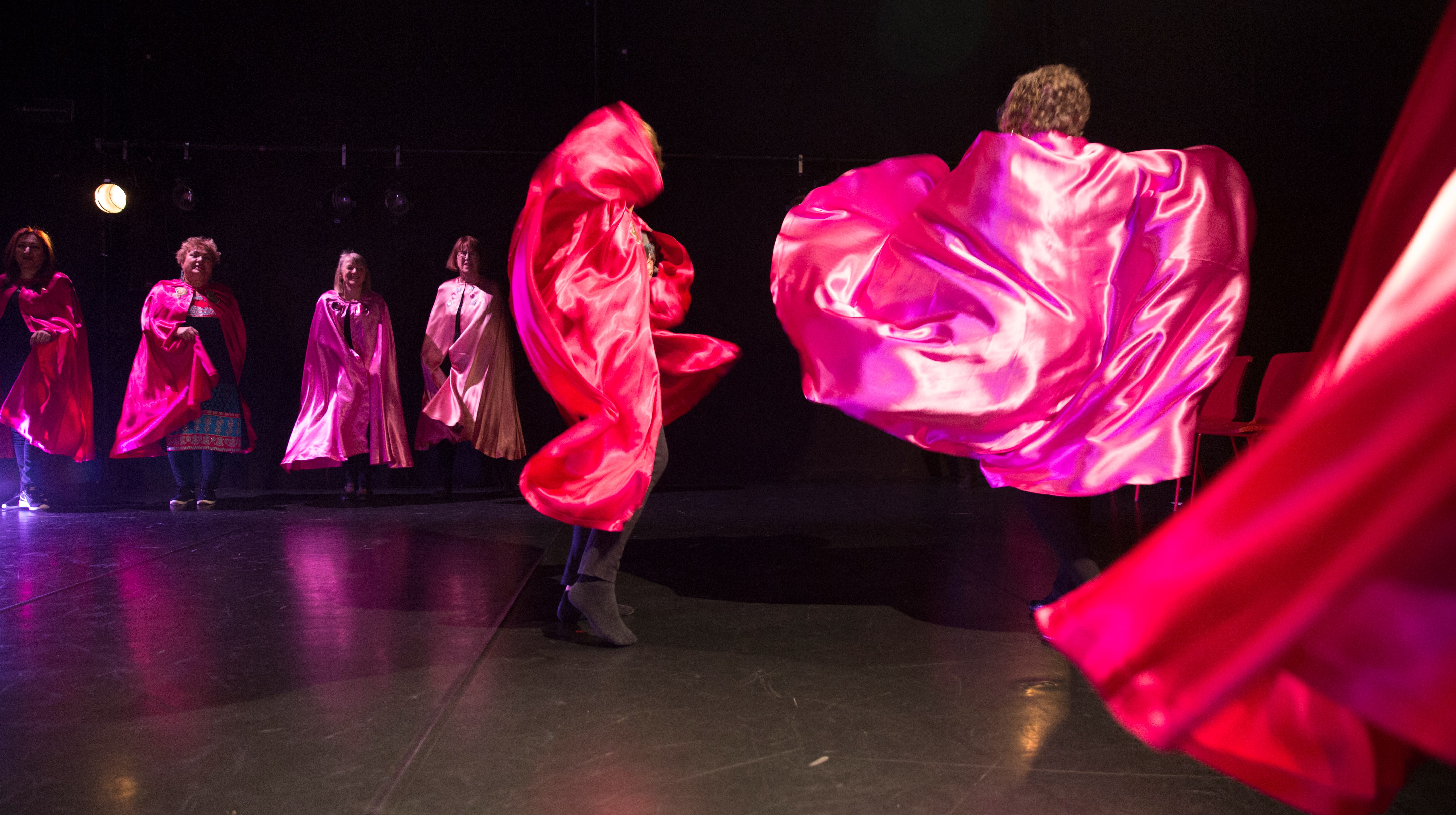 En grupp dansande kvinnor i rosa, skimrande caper, på ett scengolv.