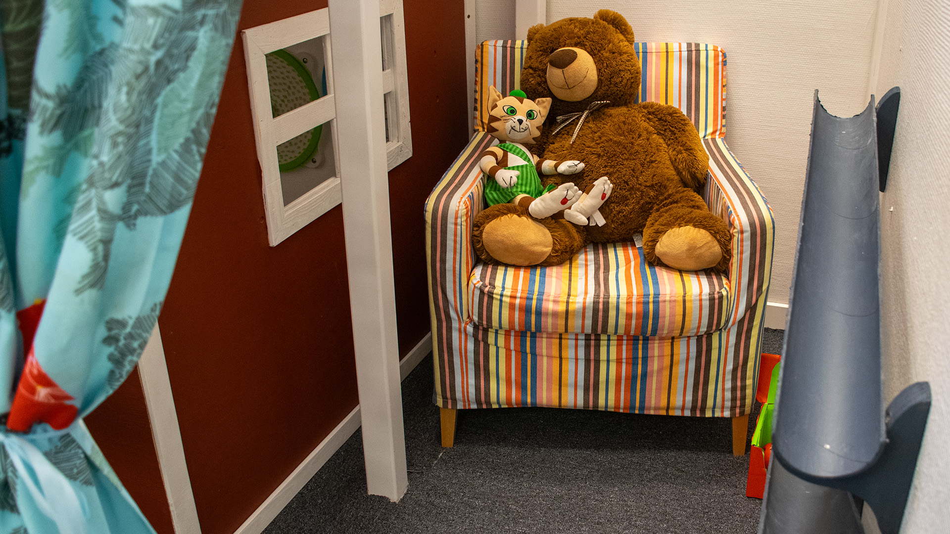En fåtölj där en stor nalle och katten Findus sitter. Fåtöljen står i ett litet hörn, på ena sidan syns snickerboas röda vägg, på andra sidan två rör på väggen som man kan rulla bollar i. 