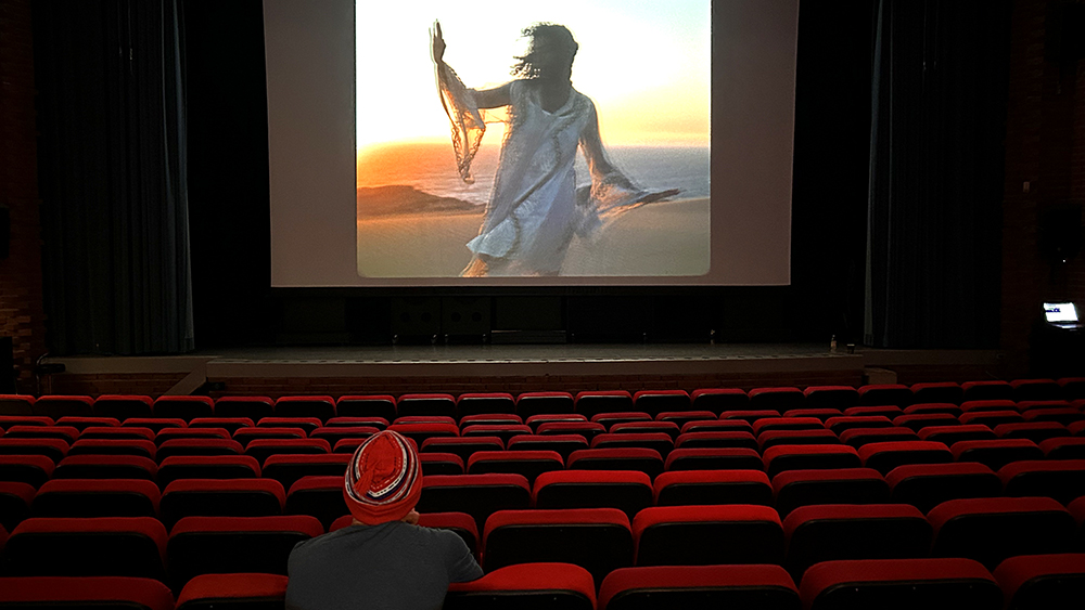 En person sitter och tittar på en bioduk, hen har en röd mössa med vita och blå streck. På bioduken syns en kvinna på en strand.