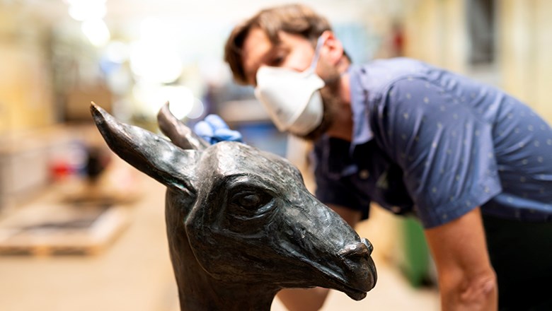 I förgrunden huvudet på en lamai metall, i bakgrunden står konservatorn, med munskydd, och penslar på laman.